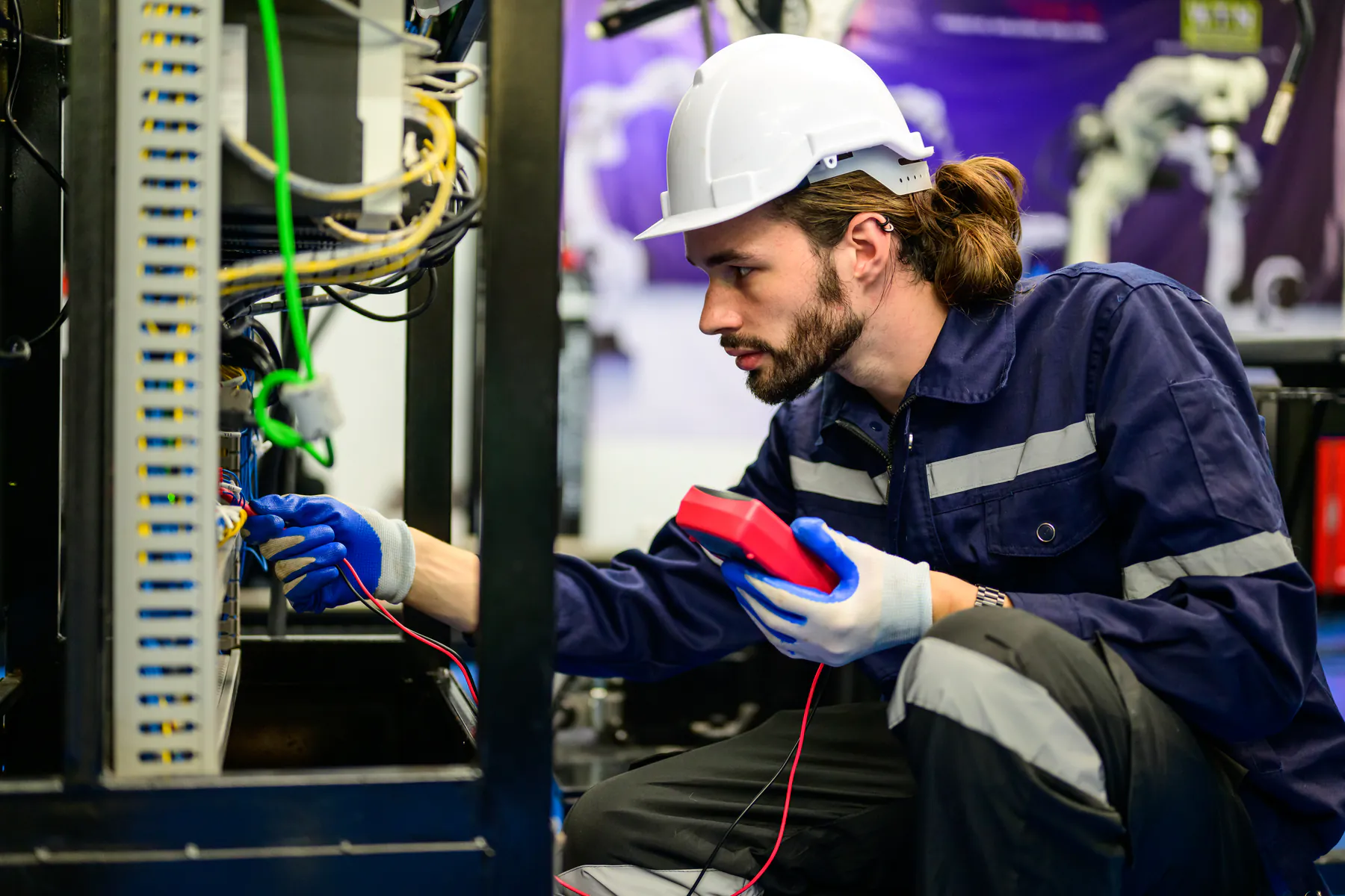 Electrician in a hard hat testing control cabinet wiring with a multimeter