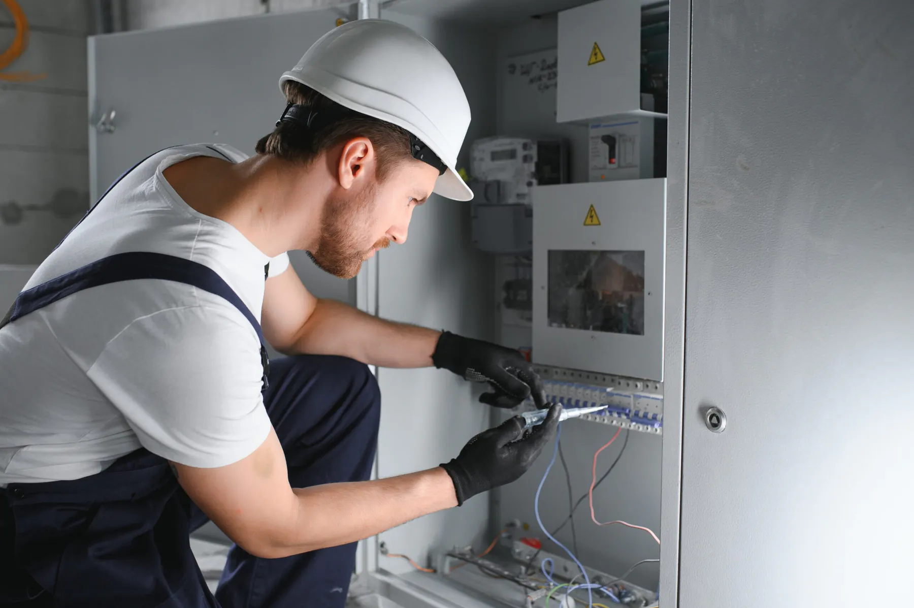 Electrician in a hard hat checking circuit breakers in a switchboard with a voltage tester